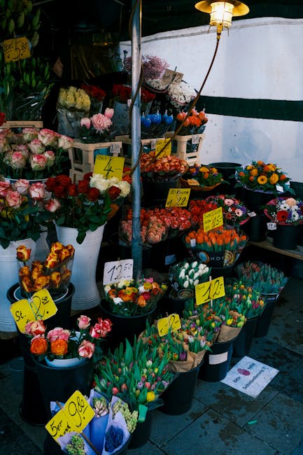 A display of various fresh flowers arranged in black buckets and white vases at a flower stall, showcasing an assortment of colourful blooms including pink roses, red tulips, orange and yellow chrysan