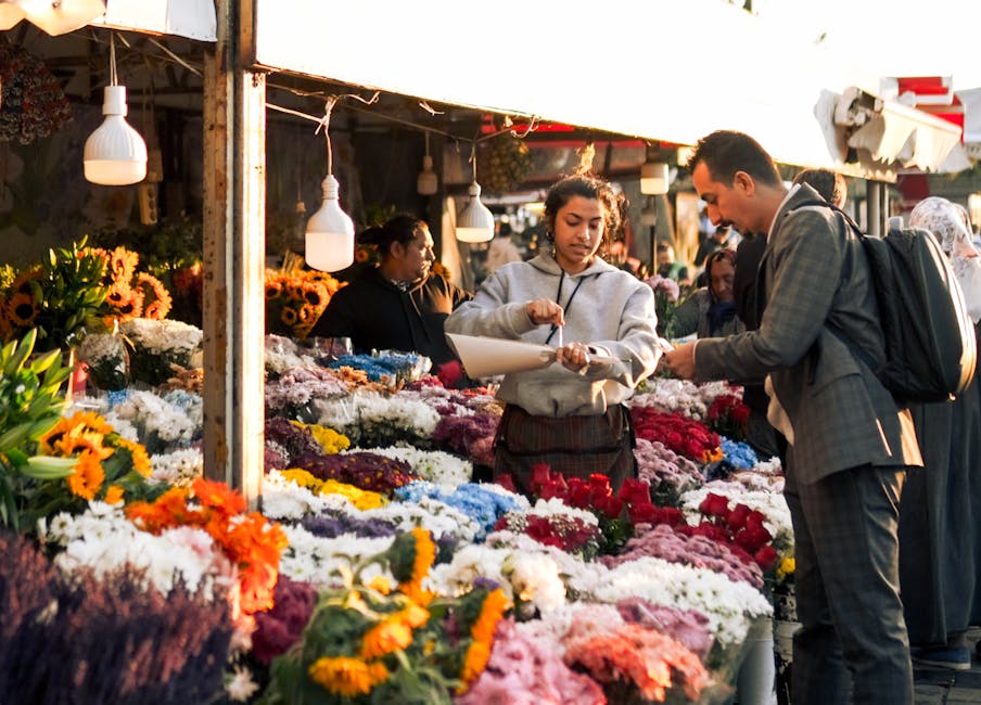 A scene at a bustling outdoor flower market illuminated by warm sunlight, featuring a wide array of vibrant flowers including sunflowers, white chrysanthemums, pink roses, purple alliums, and orange m