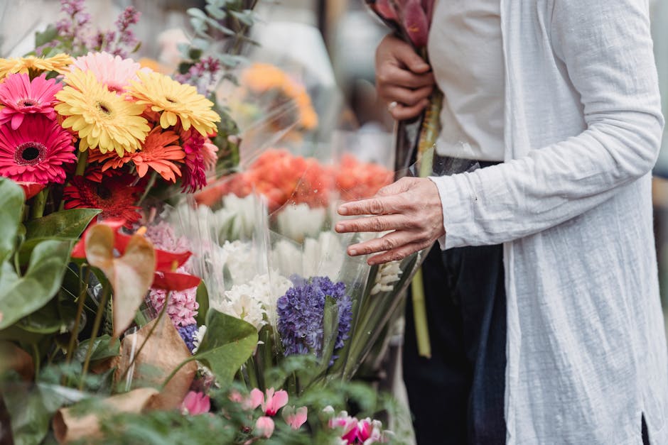 A person wearing a light grey long cardigan and dark trousers is seen at a flower market, selecting a bouquet of fresh, vibrant flowers wrapped in clear plastic. The arrangement features bright pink a