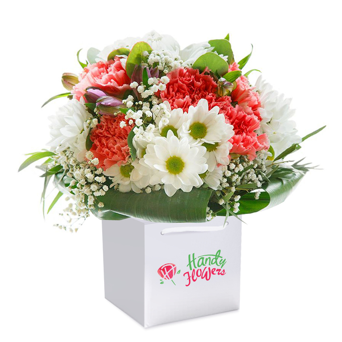 Evoking the lively energy of Earl's Court, this bouquet is presented upright in a modern white gift bag, carefully lit to showcase every vivid detail. A spirited mix of coral and peach carnations forms the heart, their abundant, ruffled petals delivering soft volume and inviting texture. Radiant white daisies with zesty yellow-green centers provide a clean, joyful accent, reminiscent of blooming neighbourhood gardens after a spring rain. Wisps of baby's breath float gracefully among the flowers, adding an airy, gentle layer, while subtle hints of unopened purple buds deepen the palette and hint at growth and renewal. Surrounding the arrangement, deep green, glossy foliage fans out in broad, elegant arcs, cradling the bouquet and providing structure, much like tree-lined avenues through the heart of Kensington and Chelsea. The crisp white gift bag-with its sturdy ribbon handles-suggests thoughtful presentation and easy transport, ready to brighten a doorstep or grace a celebration in Earl's Court. Each stem and bloom has been meticulously placed, evidence of an experienced florist's touch, all culminating in a bouquet that captures freshness, artistry, and heartfelt sentiment, ideal for birthdays, anniversaries, or everyday appreciation in SW5.