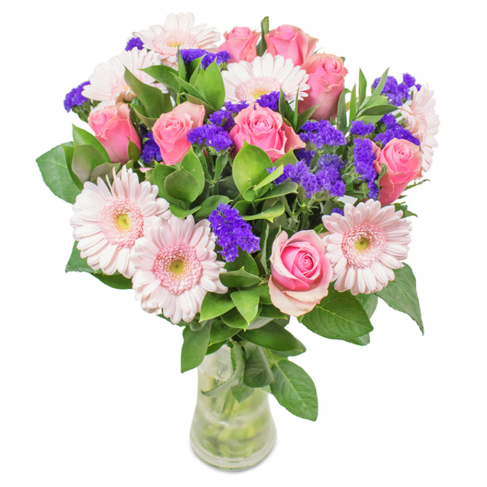 A vibrant floral arrangement sits elegantly in a clear glass vase, against a crisp white background. At its heart, you find about seven to nine medium pink roses, their petals displaying varied openness - some tightly furled and others just opening into classic spirals. These are framed beautifully by five to seven playful pale pink Gerbera daisies, their round heads wide and petals extending outwards in soft, even layers, the centers delicately pale - nearly white. Dotting the arrangement, bright purple statice introduces textural and chromatic contrast, its papery stems adding depth and interest. Deep green, glossy foliage provides a lush underpinning, filling out the bouquet with freshness familiar to those who frequent Bramham Gardens in Earl's Court. The vase's transparency frames strong stems beneath the blossoms, all bathed in clear water for a look that's as fresh as if just picked that morning. Suitable for birthdays, anniversaries, or simply brightening a light-filled flat near Old Brompton Road, this bouquet's romantic colour palette and refined structure evoke a mood of affection, celebration, and West London charm.